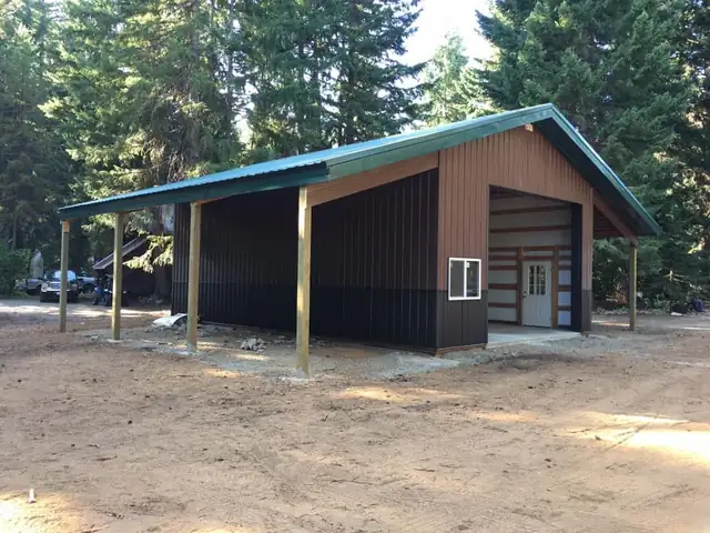 Brown and black pole building with a green metal roof, a lean-to cover and a white man door among tall pines